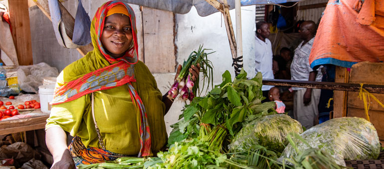 Woman in the market wearing green and orange scarf, smiling while holding onions