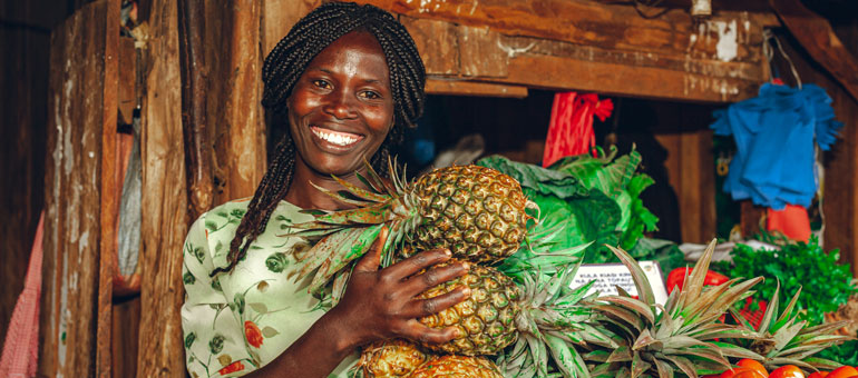 Woman holding pineapples standing next to a fruits and vegetables kiosk