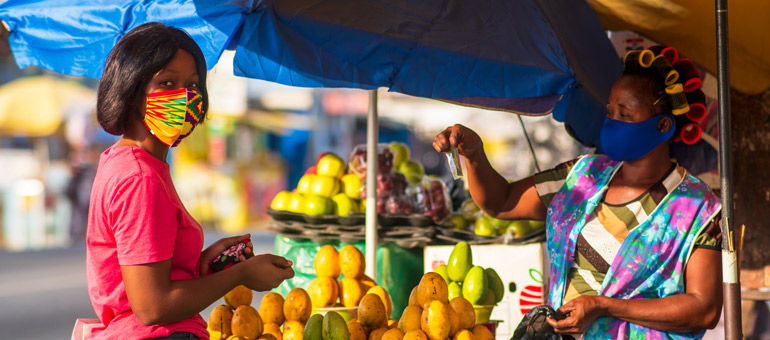 Woman buying papaya in the market from a woman in Africa both wearing colorful masks