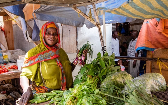 A lady holding onions in the market wearing a green dress and orange headscarf